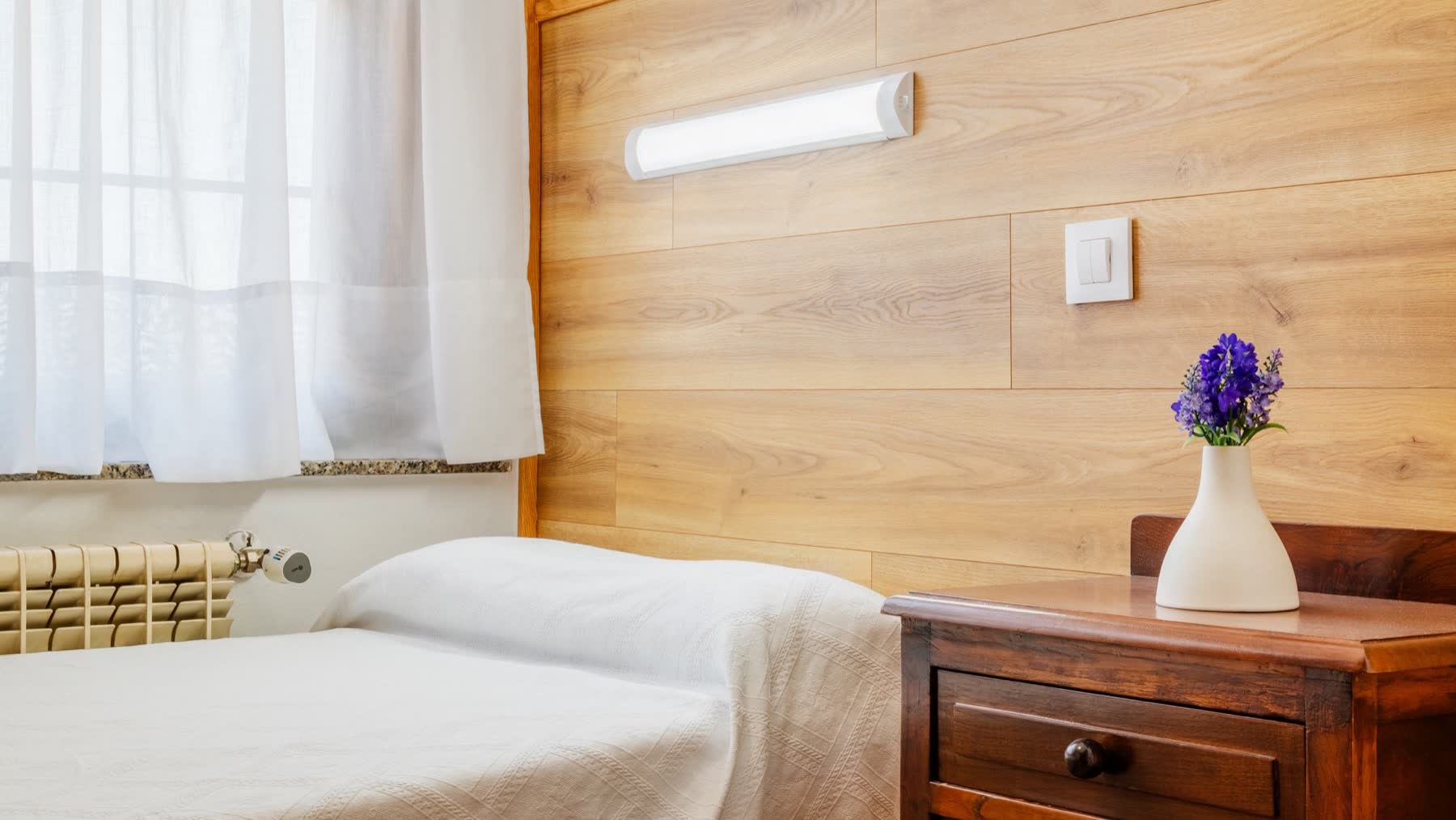 Habitación individual con cabecero de madera, mesilla con flores y ventana con visillos
