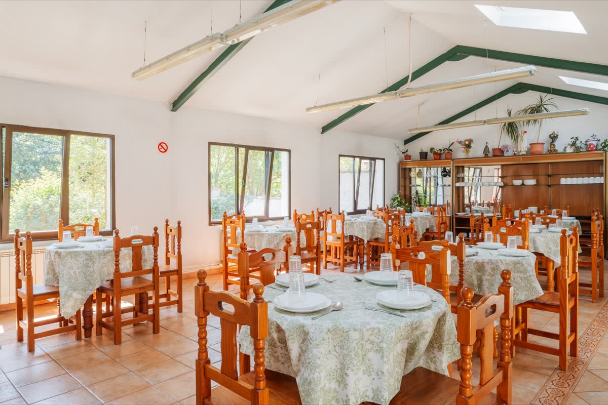 Comedor con mesas vestidas y sillas de madera, bajo una cubierta a dos aguas con ventanas al arbolado